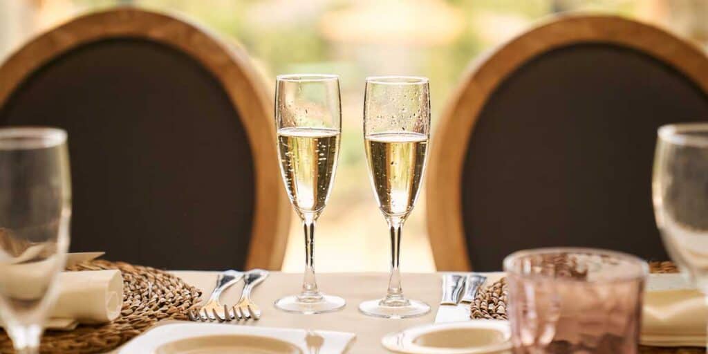 Gläser mit Sekt auf einem festlichen Esstisch, elegant dekoriert mit Geschirr und Besteck, im Hintergrund warme Holzmöbel und gemütliche Atmosphäre, geeignet für Escort Service Veranstaltungen.