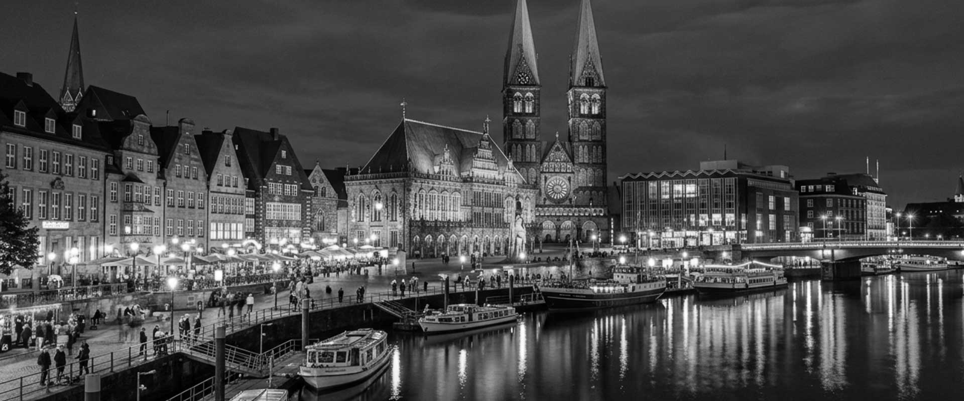 Blick auf den Hamburger Hafen mit historischen Gebäuden und Kirchen bei Nacht, beleuchtet und spiegelnd im Wasser.